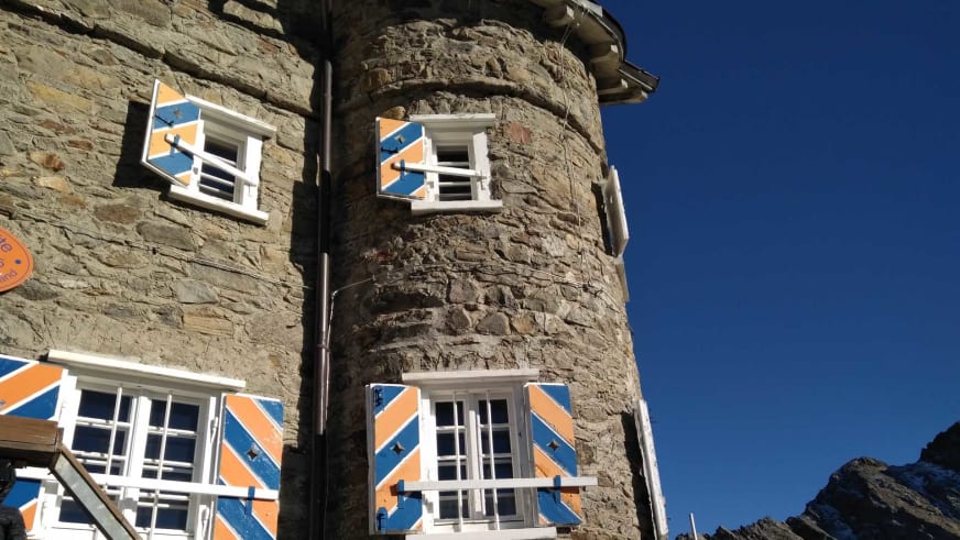 Runder Steinturm der Siegerlandhütte mit charakteristischen orange-blauen Fensterläden gegen blauen Himmel.