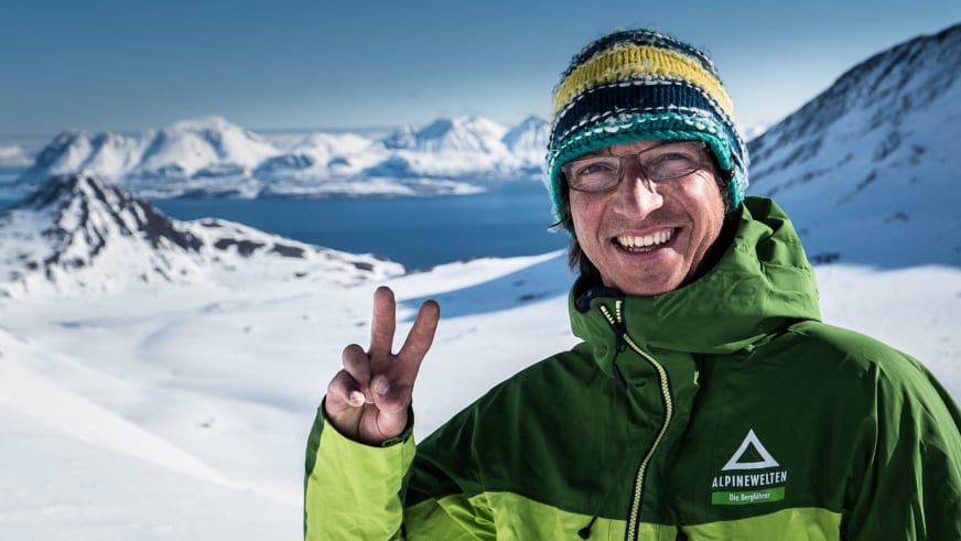 Bergführer Christoph Hummel zeigt Victory-Zeichen vor Fjordlandschaft mit schneebedeckten Bergen in Norwegen.