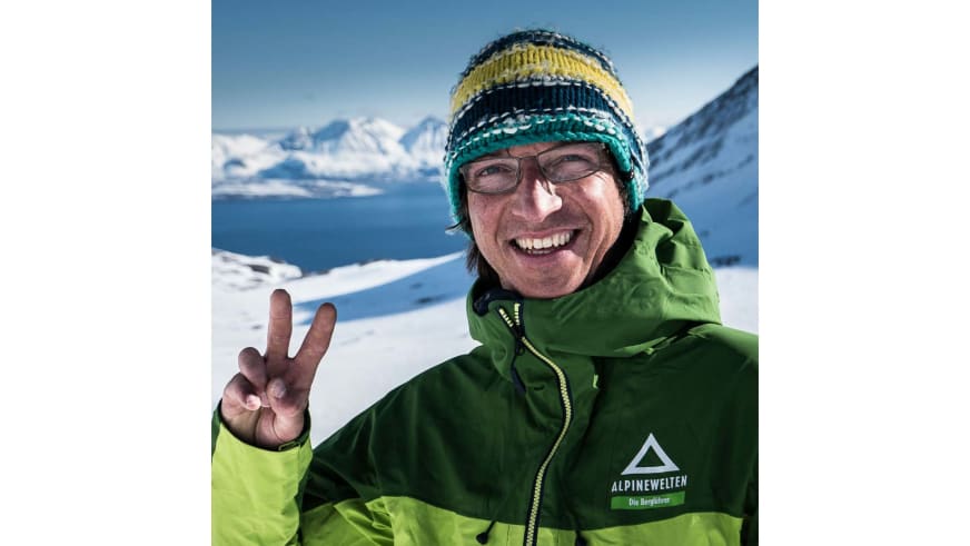 Lächelnder Bergführer Christoph Hummel zeigt Victory-Zeichen vor verschneiter Fjordlandschaft mit blauem Himmel.