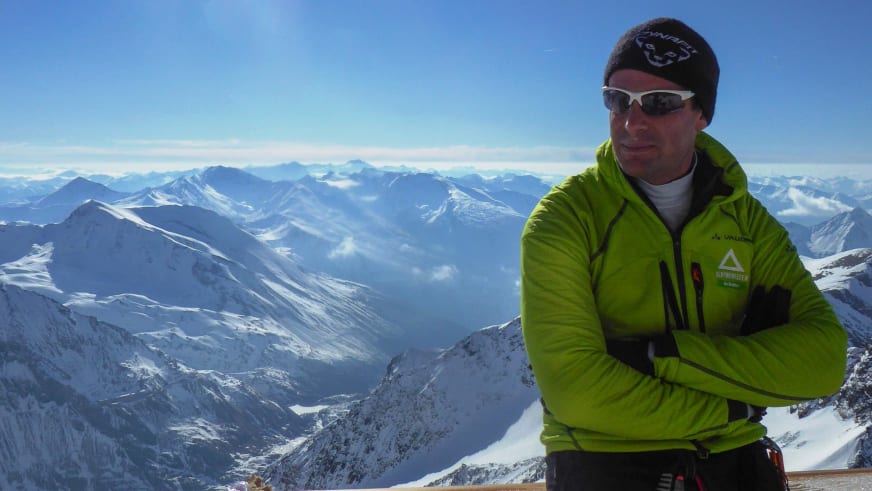 Bergführer Simon Kraus steht mit verschränkten Armen auf schneebedecktem Gipfel mit Bergpanorama im Hintergrund.