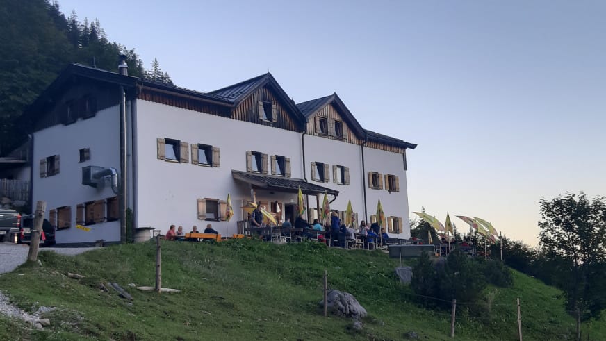 Außenansicht der Gaudeamushütte mit Terrasse und Gästen auf grüner Almwiese im Wilden Kaiser.