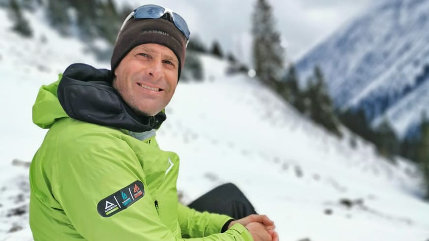 Portrait von Bergführer Simon Kraus in grüner Skijacke und schwarzer Mütze vor verschneiter Berglandschaft.