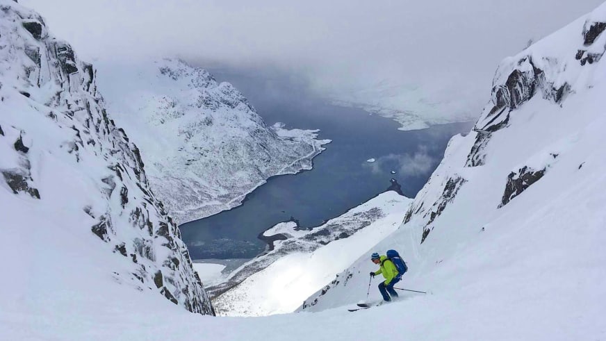 Bergführer Simon Kraus fährt auf Skiern einen steilen Hang hinab mit tiefblauem Fjord und Bergen im Hintergrund.
