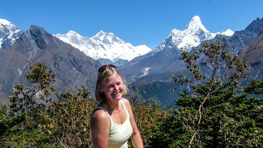 Portrait von Bergwanderführerin Isolde Fink im weißen Top vor schneebedeckten Berggipfeln und grüner Vegetation.