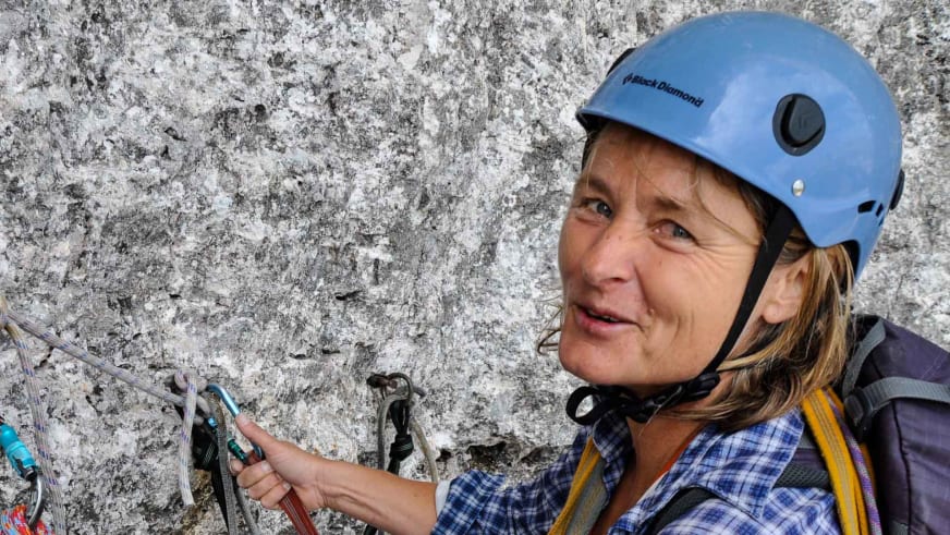 Bergwanderführerin Isolde Fink trägt blauen Kletterhelm und kariertes Hemd vor heller Felswand mit rotem Gerät in der Hand.