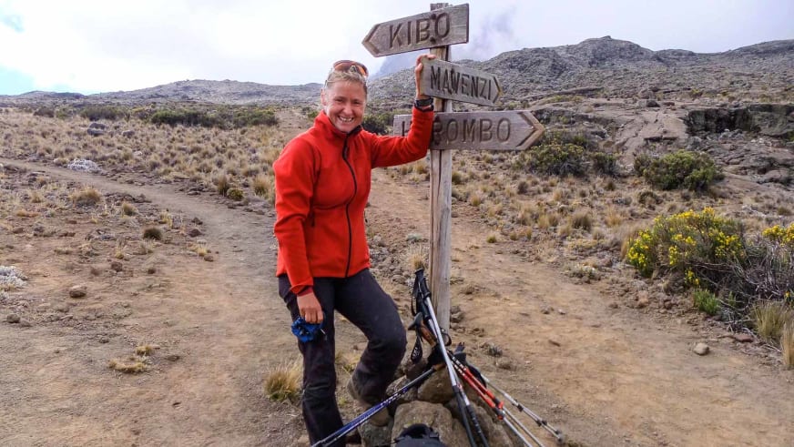 Bergwanderführerin Isolde Fink posiert lächelnd neben hölzernem Wegweiser in karger Hochebene mit Trekkingstöcken.