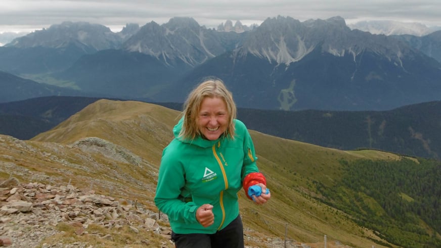 Bergwanderführerin Isolde Fink in grüner Alpine Welten Jacke lächelt auf steinigem Bergrücken vor bewölkter Kalksteinwand.