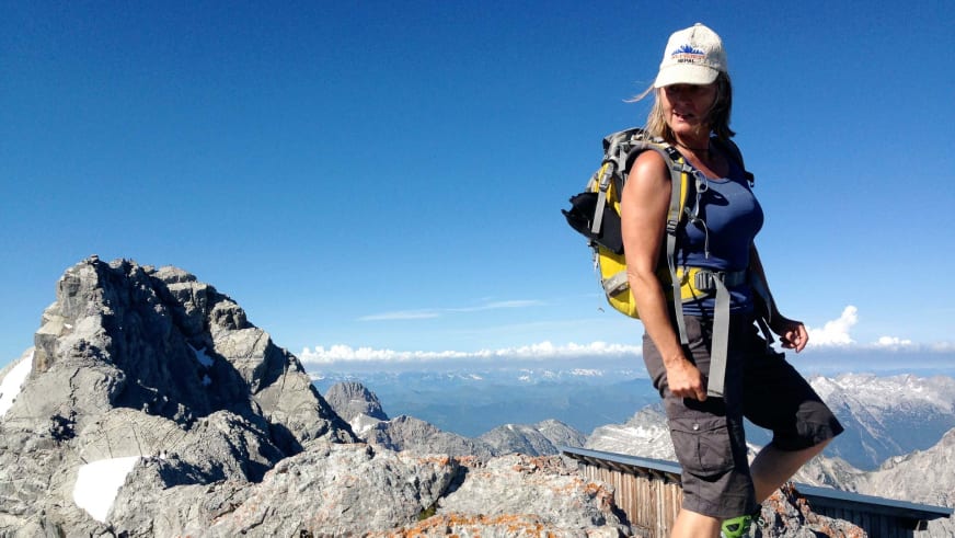 Bergwanderführerin Isolde Fink steht auf felsigem Gipfelplateau mit Wanderstock vor Bergpanorama und blauem Himmel.