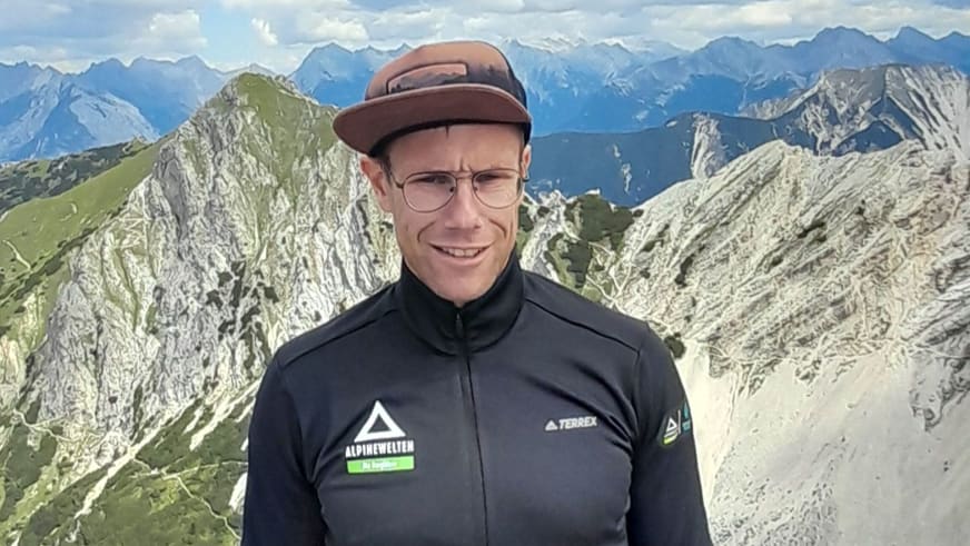 Bergführer Jens Lang lächelt in die Kamera vor einer Berglandschaft mit steilen Kalksteinwänden und grüner Vegetation.