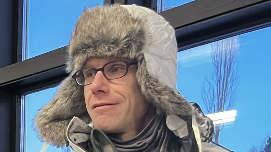Bergführer Jens Lang trägt eine graue Fellmütze und dunkle Brille in einem beheizten Innenraum vor großen Fenstern.