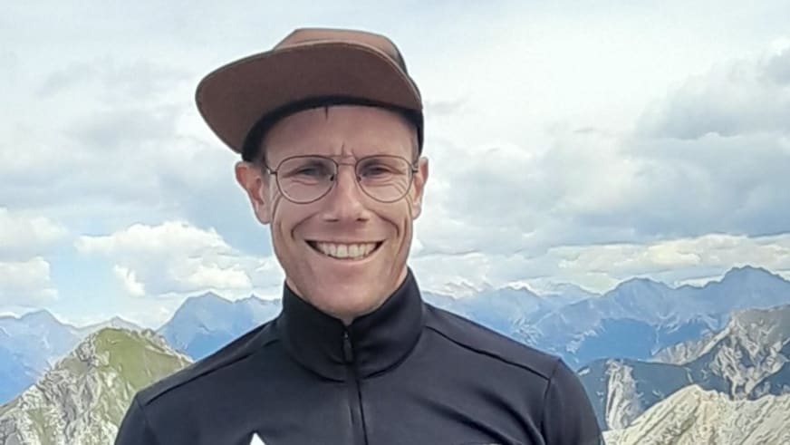 Bergführer Jens Lang trägt braune Baseballkappe und Brille vor einem Bergpanorama mit mehreren Gebirgsketten.