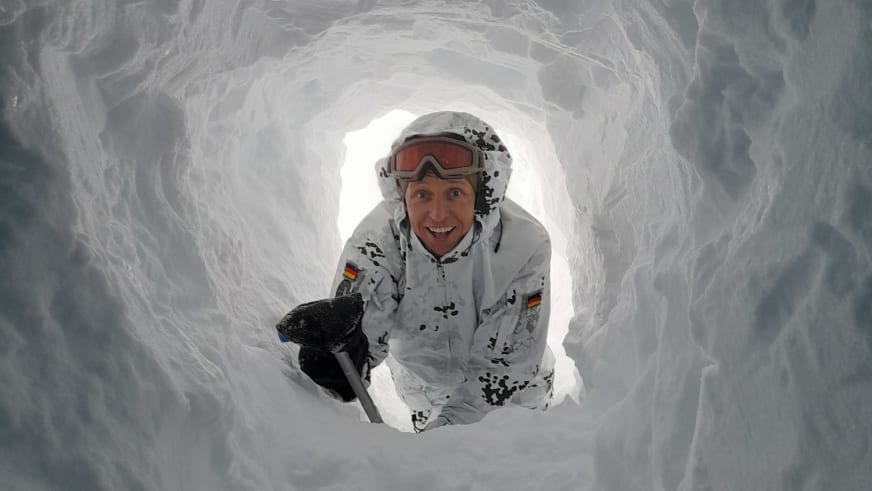 Bergführer Jens Lang blickt aus einer Schneehöhle und hält eine kleine Schaufel in der rechten Hand.