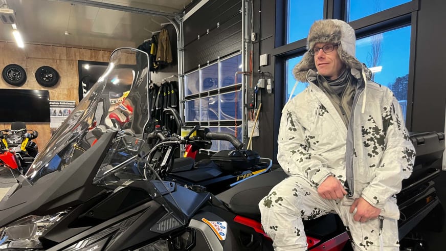 In einer Fahrzeughalle sitzt Bergführer Jens Lang mit Fellmütze neben einem schwarzen Schneemobil.