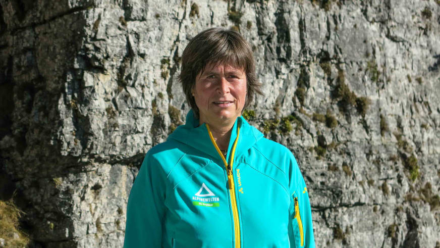 Bergwanderführerin Angelika Neubauer lächelt vor einer hellen Kalksteinwand und trägt eine türkise Softshell-Jacke.