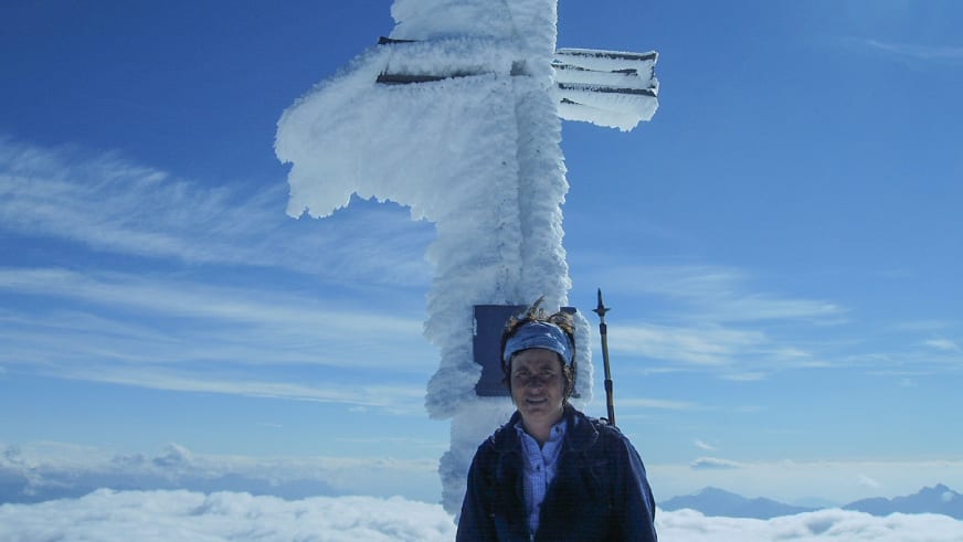 Bergwanderführerin Angelika Neubauer steht vor einem stark vereisten Gipfelkreuz mit bizarren Eisformationen bei blauem Himmel.