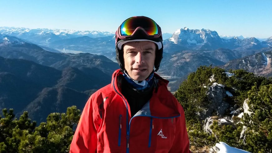 Bergführer Jörg Rauschenberger trägt rote Funktionsjacke mit Skibrille auf der Stirn vor schneebedeckten Bergen.