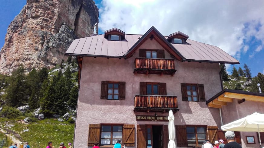 Wanderer vor dem Rifugio Cinque Torri, das unterhalb einer steilen Kalksteinwand in den Dolomiten auf Almwiesen steht.