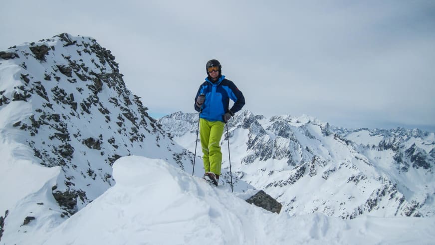 Auf einem verschneiten Berggipfel steht Bergwanderführer Karlheinz Rösch mit Skistöcken vor Bergpanorama.