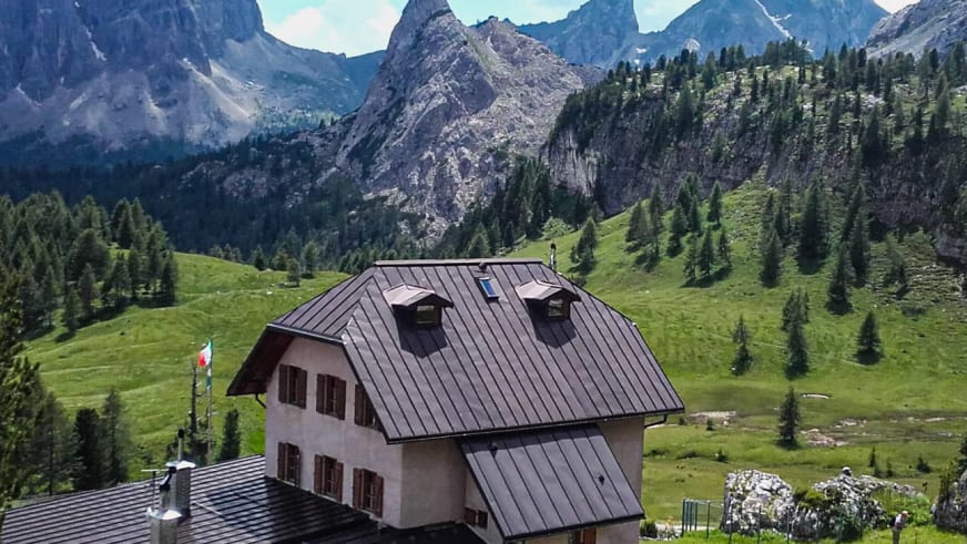 Das Rifugio Cinque Torri steht auf grüner Almwiese vor steilen Kalksteinwänden in den italienischen Dolomiten.