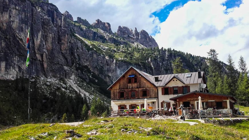 Das Rifugio Croda da Lago liegt auf grüner Almwiese mit großer Terrasse vor steiler Dolomiten-Felswand.