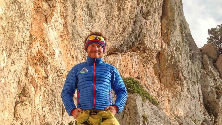 Bergführer Marcel Roßbach lächelt in blauer Daunenjacke vor einer hohen gelblich-grauen Felswand.