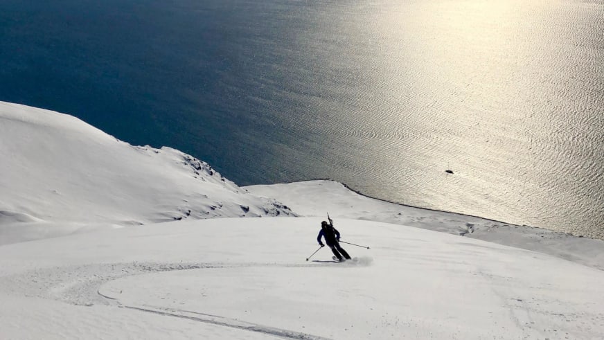 Bergführer Marcel Roßbach fährt auf Skiern einen verschneiten Hang hinab mit goldenem Fjord im Hintergrund.