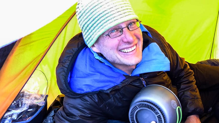 Im gelben Zelt hält Bergführer Michael Rüter ein technisches Gerät und trägt gestreifte Mütze mit blauer Daunenjacke.