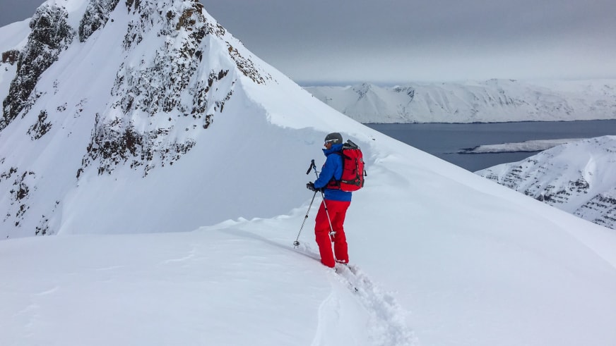 Bergführer Florian Schmidt in roter Hose und blauer Jacke steigt einen verschneiten Hang in isländischer Fjordlandschaft auf.
