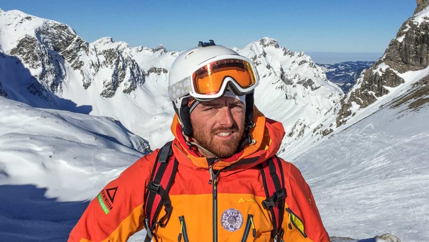 Portrait von Bergführer Florian Schmidt in oranger Daunenjacke mit weißem Skihelm vor verschneiter Bergkulisse.