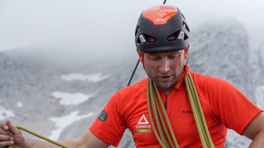 Guide Markus Schubert mit orangem Petzl-Kletterhelm hält gelbes Kletterseil vor Berglandschaft mit grauen Felswänden.