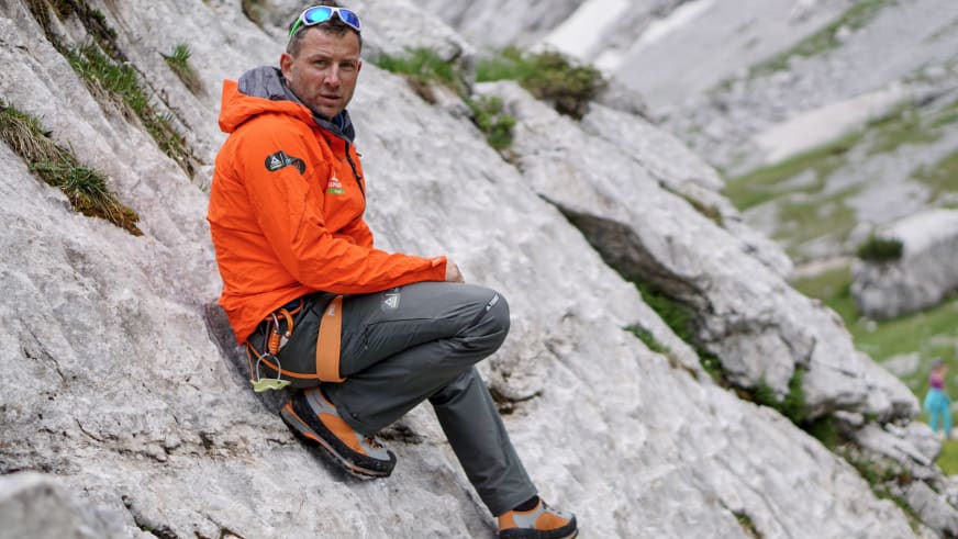 Guide Markus Schubert sitzt in orangefarbener Outdoorjacke auf hellem Kalksteinfels mit Klettergurt und Bergschuhen.