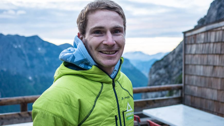 Bergführer Christoph Sokoll lächelt auf einer Holzterrasse in grüner Vaude Daunenjacke mit Berglandschaft im Hintergrund.