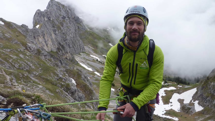 Bergführer Philipp Berg trägt weißen Kletterhelm und schwarzen Klettergurt vor alpiner Berglandschaft mit Kalksteinfelsen.