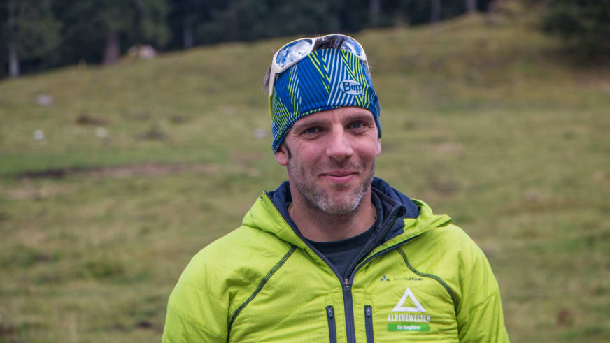 Auf einer grünen Almwiese posiert Bergführer Andreas Wiedenmann mit grüner Jacke und Stirnlampe an der Mütze.