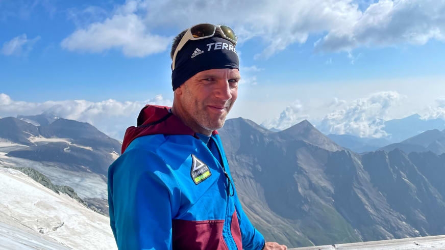 Portrait von Andreas Wiedenmann, Bergführer mit blauer Softshell-Jacke und Sonnenbrille vor schneebedeckter Bergkulisse.