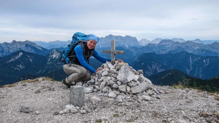 Bergwanderführerin Sandra Wolf kniet neben einem Steinmännchen mit Holzkreuz auf einem Berggipfel und lächelt in die Kamera.