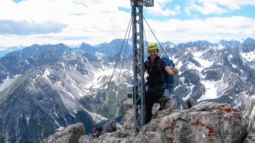 Bergwanderführerin Sandra Wolf steht lächelnd am Metallkreuz eines hohen Berggipfels mit schneebedeckten Alpengipfeln im Hintergrund.