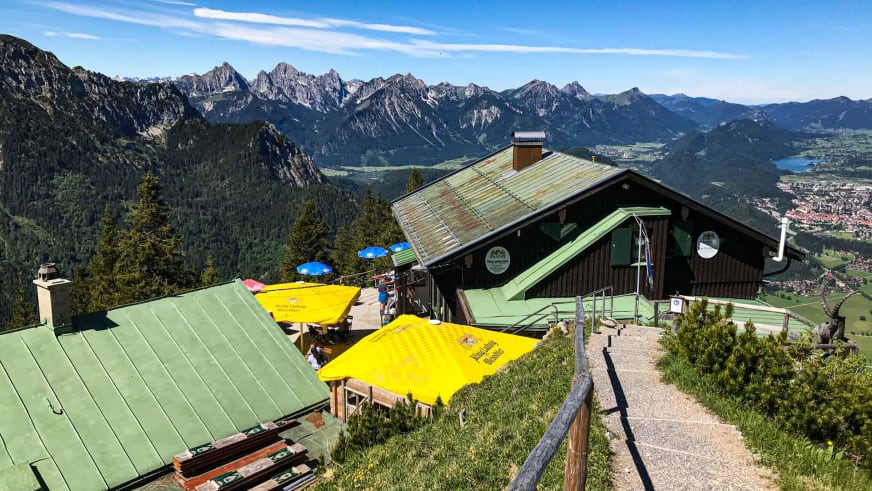 Panorama des Tegelberghaus mit Sonnenterrasse und Blick über die bewaldeten Täler der Ammergauer Alpen.