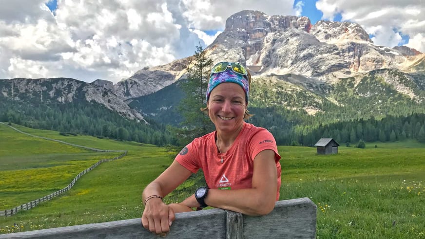 Bergwanderführerin Sandra Wolf stützt sich lächelnd auf einen Holzzaun und trägt ein rotes T-Shirt mit Alpine Welten Logo.