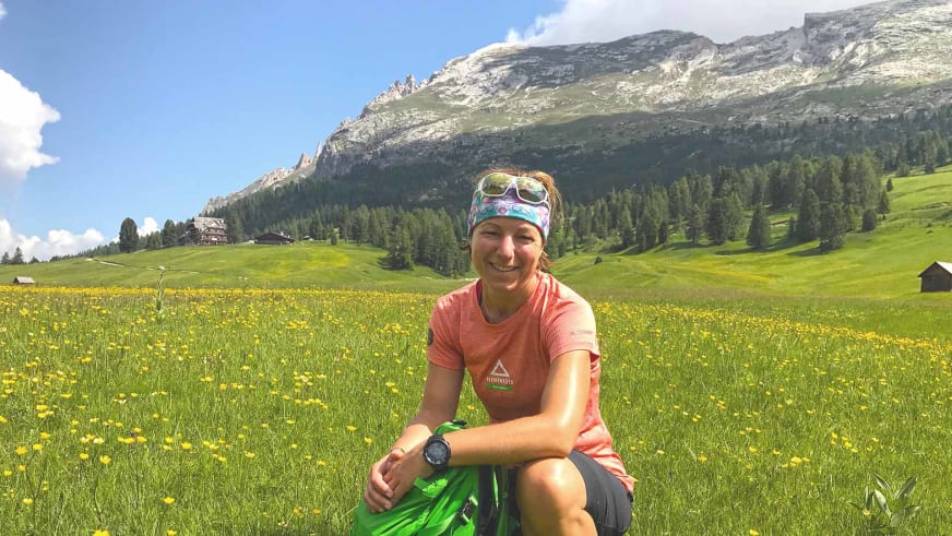 Bergwanderführerin Sandra Wolf sitzt lächelnd auf einer Blumenwiese mit gelbem Stirnband und grünem Rucksack neben sich.