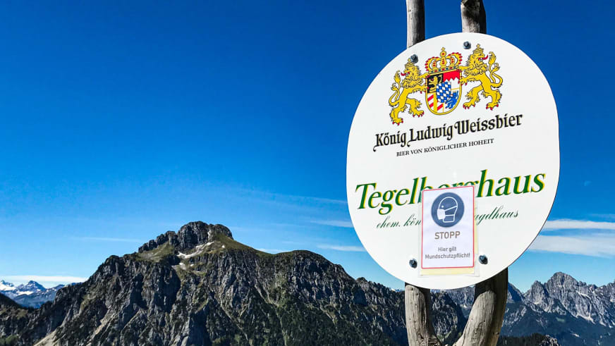 Rundes Hüttenschild mit bayerischem Wappen am Tegelberghaus vor Bergpanorama der Ammergauer Alpen.
