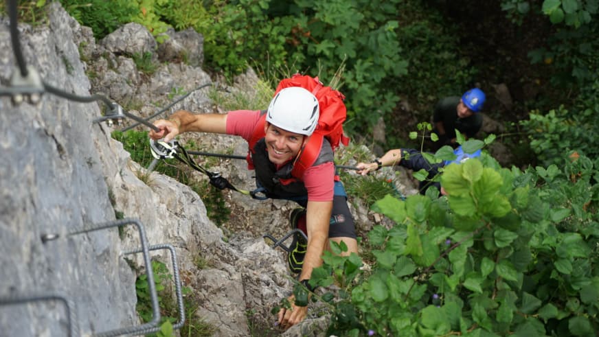 Bergführer Christian Knabe klettert am Drahtseil einer Felswand in roter Jacke und weißem Helm mit Klettersteig-Set.