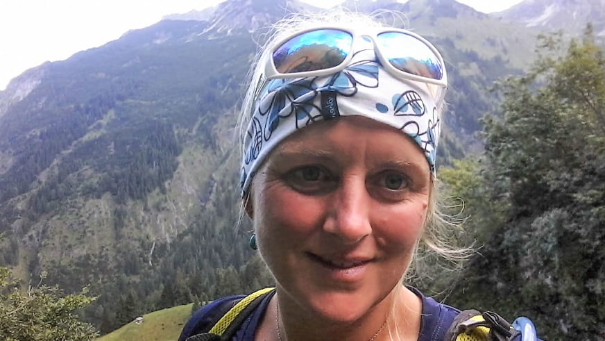 Bergwanderführerin Maria Anna Huber lächelt mit weißer Gletscherbrille und blauem Multifunktionstuch vor bewaldeter Berglandschaft.