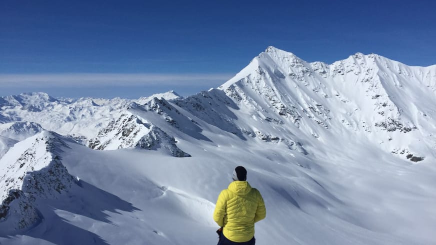 Bergführer Rene Hellmann in gelber Daunenjacke betrachtet schneebedeckte Gipfel am Elbrus bei strahlendem Himmel.
