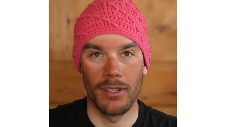 Bergführer Philipp Neumann trägt eine rosa gestrickte Wollmütze und schwarzes Shirt in einem Innenraum mit Holzwand.