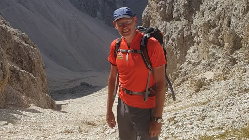 Bergwanderführer Manfred Beier lächelt in blauer Kappe und rotem T-Shirt mit Klettergurt vor hellen Kalksteinformationen.