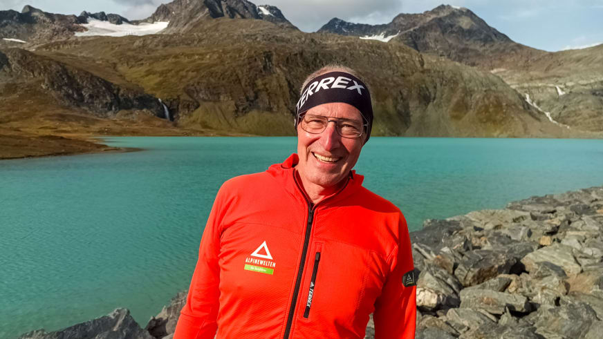 Bergwanderführer Manfred Beier in roter Softshelljacke mit Alpine Welten Logo steht am steinigen Ufer eines türkisfarbenen Bergsees in Norwegen.