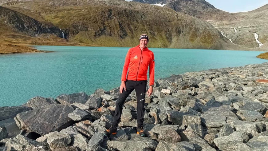 Auf Geröllfeld steht Bergwanderführer Manfred Beier in roter Outdoorjacke vor türkisblauem Bergsee in norwegischer Berglandschaft.