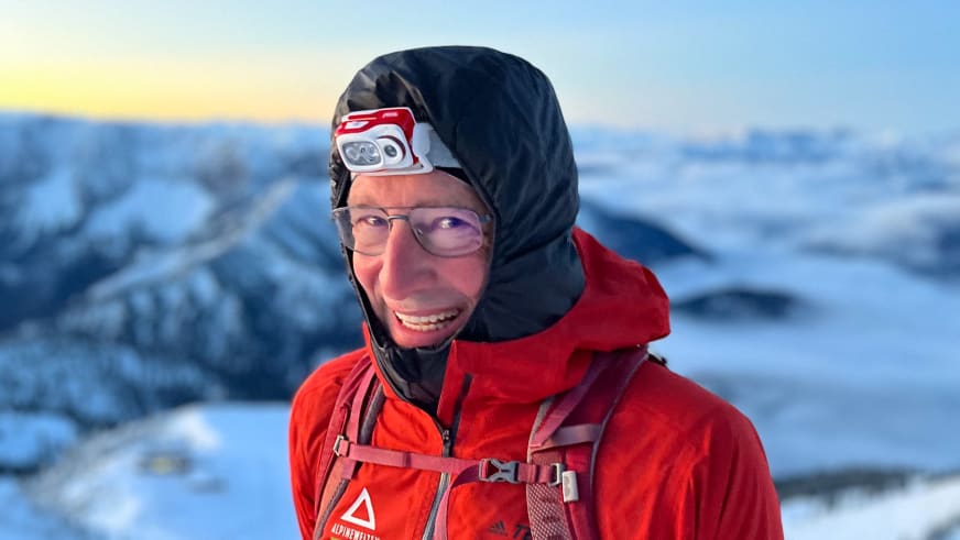 Bergwanderführer Manfred Beier trägt rote Winterjacke, schwarze Kapuze und Stirnlampe bei goldenem Licht in schneebedeckter Berglandschaft.