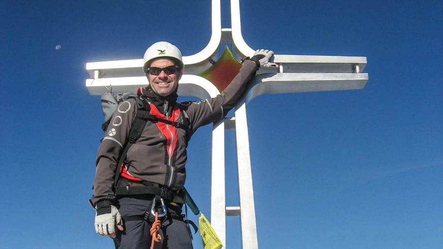 Portrait von Bergwanderführer Andreas Schneider mit weißem Helm und Sonnenbrille vor einem weißen Gipfelkreuz unter blauem Himmel.